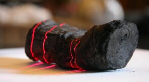 Zwęglony, zwinięty papirus leżący na stole podczas skanowania; na jego powierzchni widoczne są czerwone linie lasera.