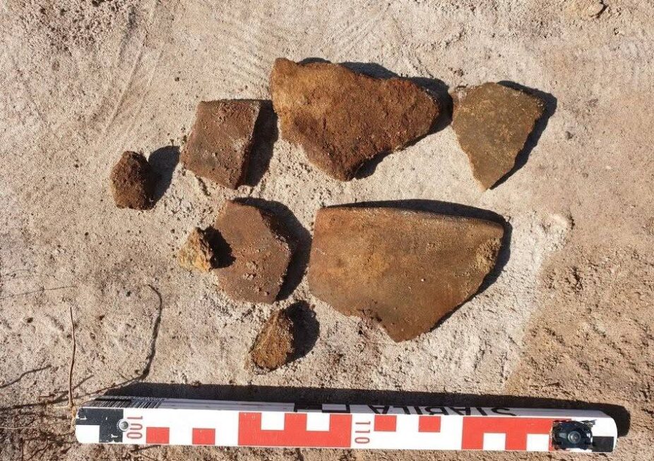 Kilka fragmentów pradziejowych naczyń ceramicznych ułożonych na jasnym piasku obok skali archeologicznej.