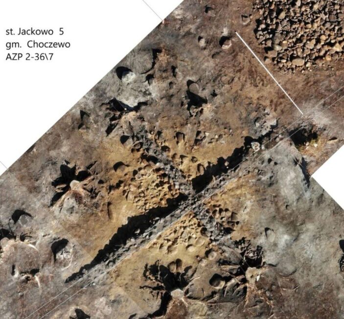 Widok z góry na wykop archeologiczny w Jackowie z widocznymi skupiskami kamieni i ciemniejszymi śladami obiektów w ziemi.