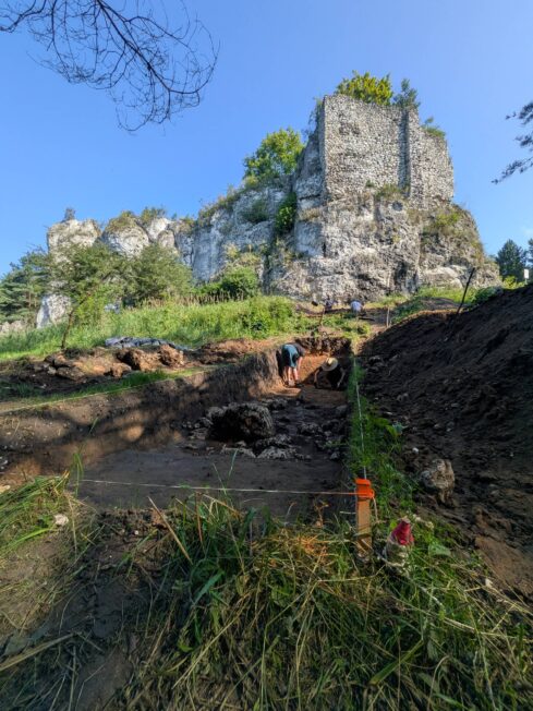 Archeolodzy pracujący w wykopie, nad którym wznosi się wapienny ostaniec z zachowaną ścianą średniowiecznej strażnicy.