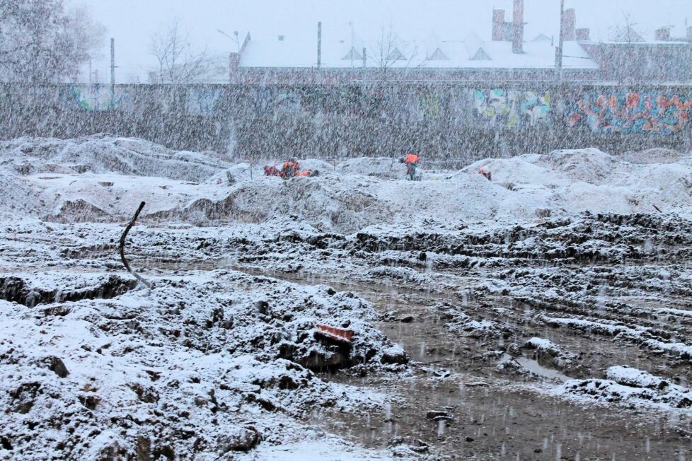 Rozległe wykopaliska archeologiczne podczas intensywnych opadów śniegu, z kałużami, hałdami ziemi i archeologami pracującymi w kamizelkach odblaskowych