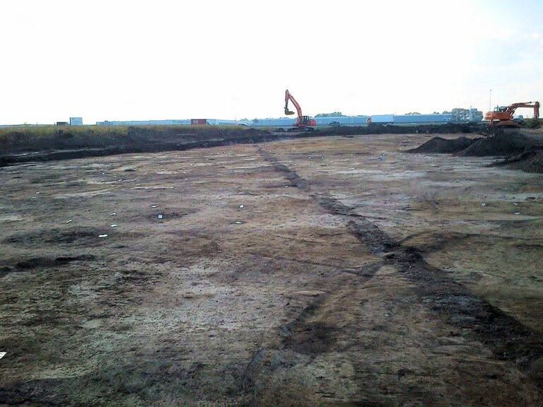 Rozległa, wyrównana powierzchnia stanowiska archeologicznego po zdjęciu nadkładu, z koparkami pracującymi w oddali