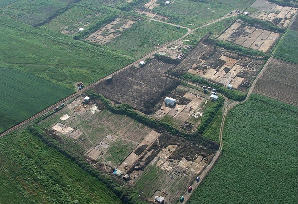 Szeroki, ukośny kadr lotniczy; wśród zielonych pól kilka dużych prostokątnych obszarów wykopów archeologicznych z siatką wykopów i drogami dojazdowymi