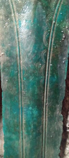 Zbliżenie zielonkawej głowni brązowego miecza z rytym ornamentem z łuków i krótkich kresek.