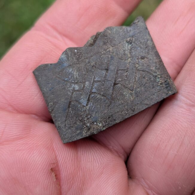Fragment ciemnego metalowego okucia z rytym ornamentem geometrycznym, trzymany na dło