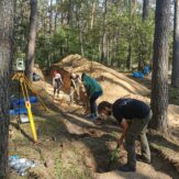 Zespół badawczy prowadzi prace ziemne przy wykopie archeologicznym w lesie, obok ustawiony jest tachimetr.