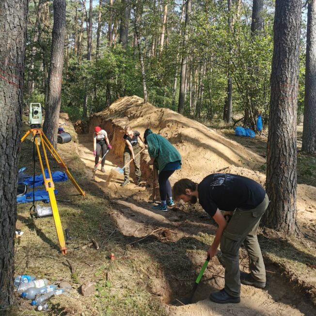 Zespół badawczy prowadzi prace ziemne przy wykopie archeologicznym w lesie, obok ustawiony jest tachimetr.