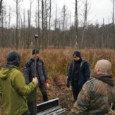 Badacze stoją na torfowisku z wysoką roślinnością i sprzętem do pobierania rdzeni osadów